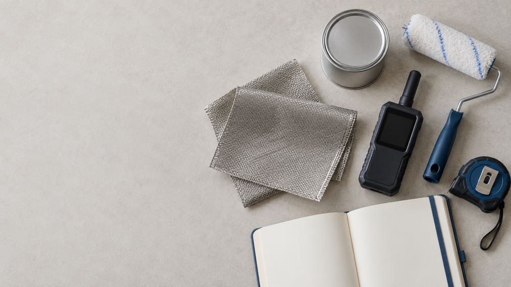 The pre-purchase checklist, laid out: materials, meter, and a notebook for the numbers A flat-lay of EMF shielding materials — a conductive mesh swatch, a paint sample tin, a roller, a handheld RF meter, a tape measure, and an open blank notebook — on a pale tabletop